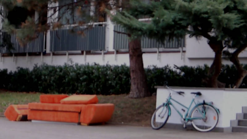 movie still shows a scene in front of a city house, an old orange couch and a blue bicycle under some trees